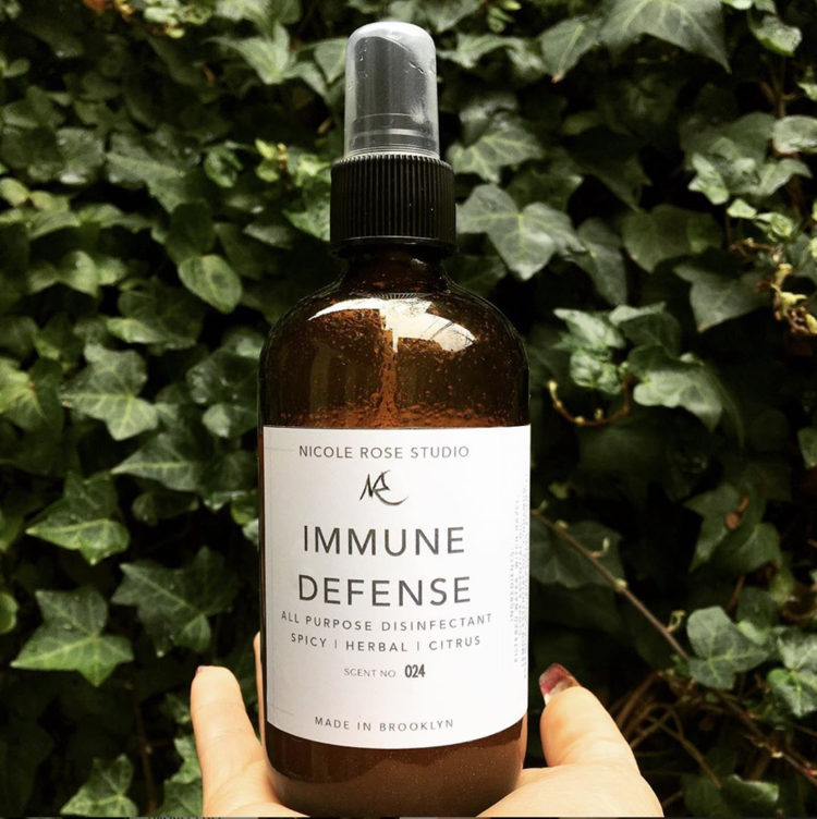 Immune Defense all-purpose spray in a 4 oz amber glass bottle, held against a wall of ivy
